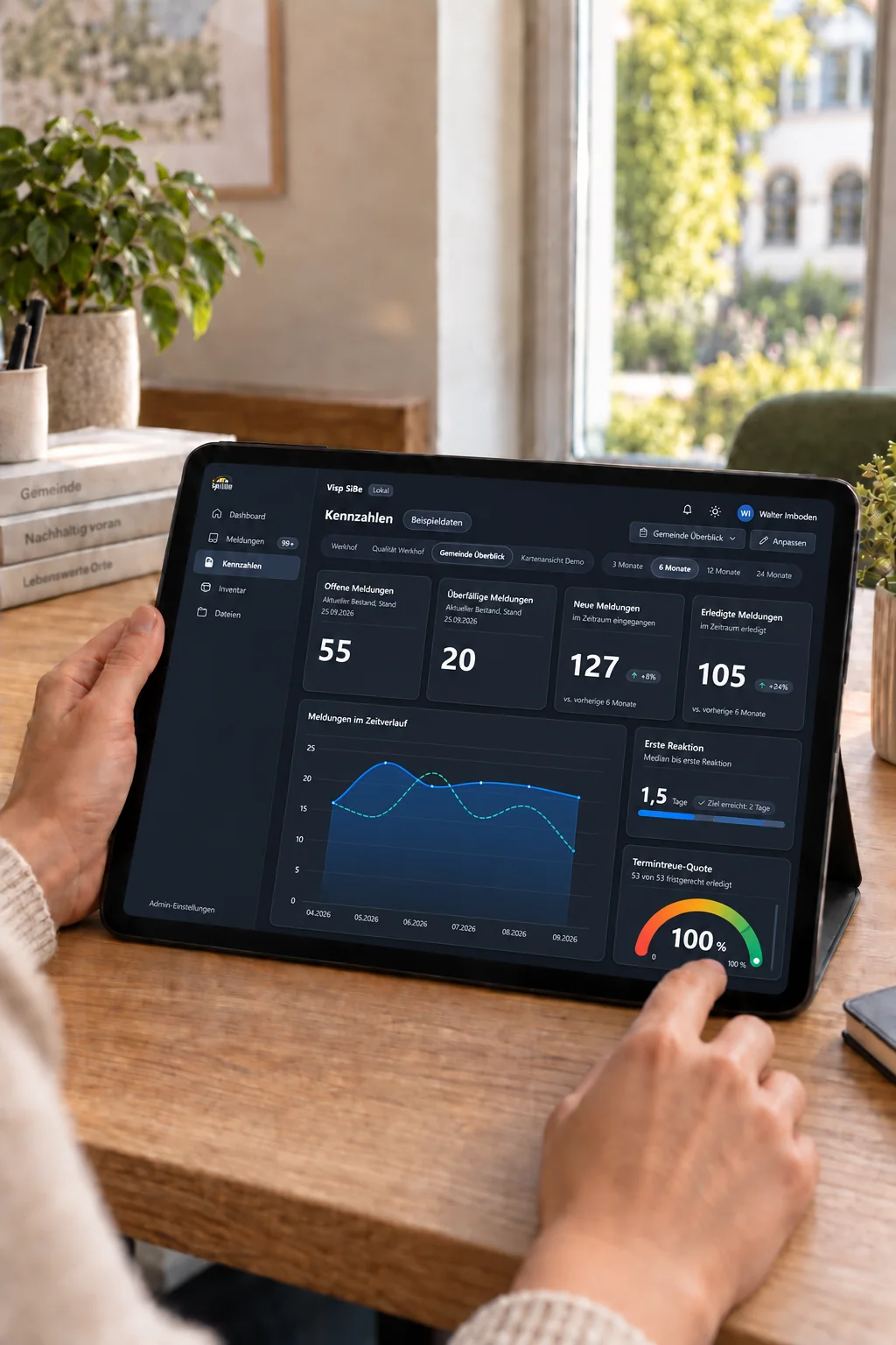Illustrative Szene im Gemeindebüro: Eine Person sieht auf einem Tablet mit dunkler Oberfläche vier als Beispieldaten gekennzeichnete Visp SiBe Kennzahlen zu Meldungen und ein Liniendiagramm.